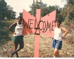 Two women holding a tall homemade pink plywood sign in the shape of a labrys, with Welcome painted on it