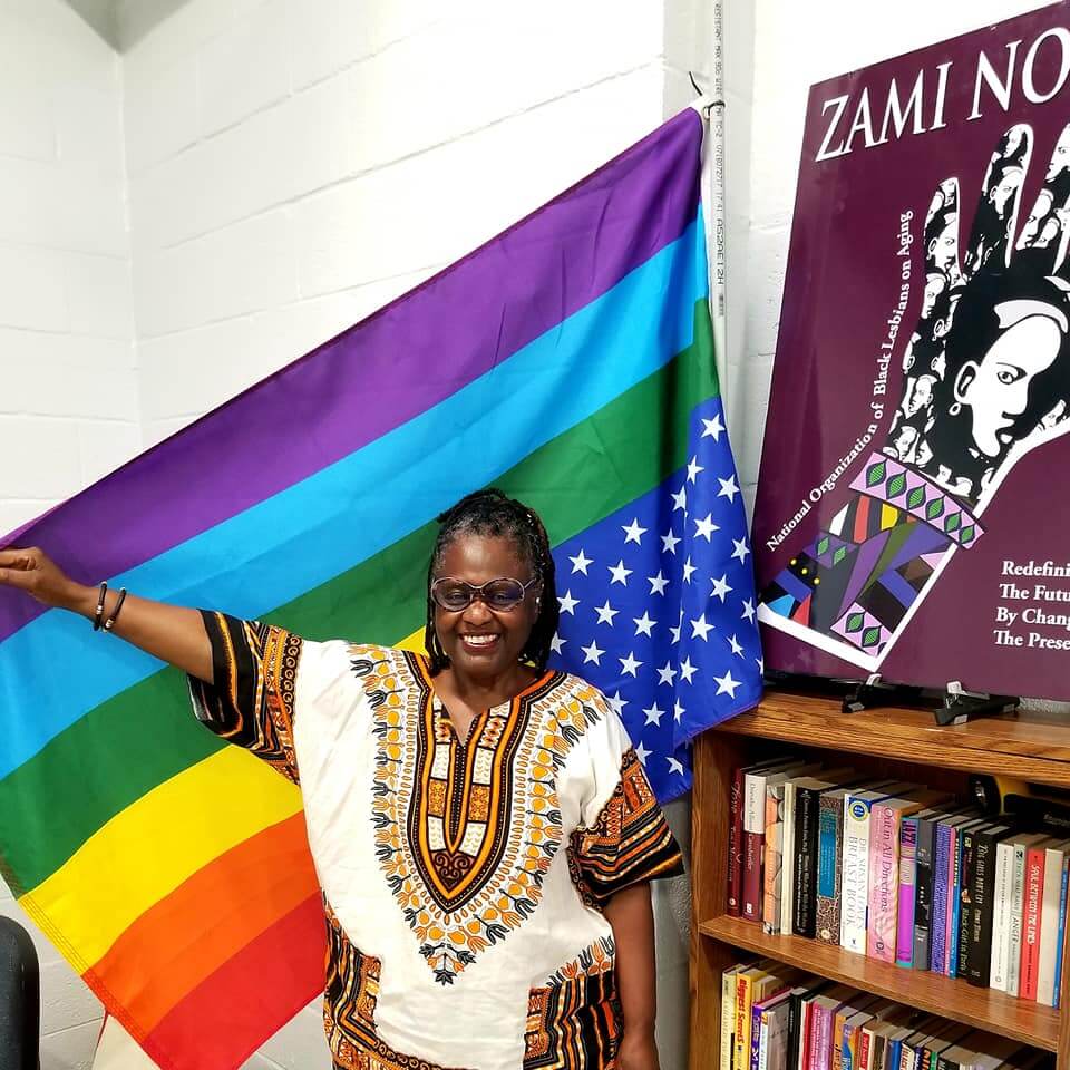 Mary Anne Adams standing n a dashiki and with one arm up against the rainbow flag