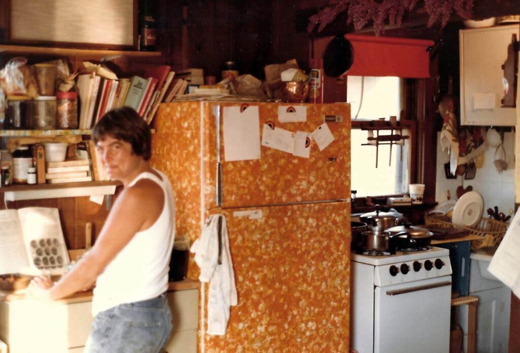 Lavender Lieu looks over her shoulder next to a speckled refrigerator in a cluttered kitchen