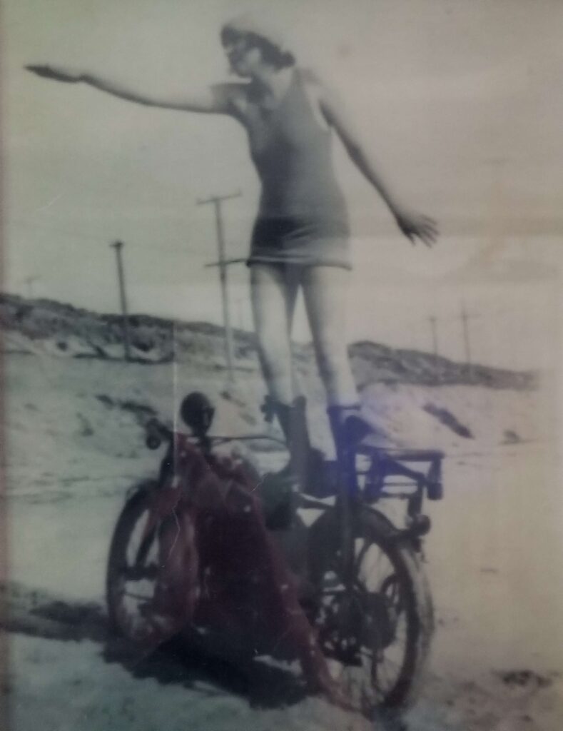 Great grandmother called "Maude with the bod", standing on the seat of a Harley Davidson motorcycle wearing a one-piece bathing suit and boots and a cap in a stance like she is surfing, on the beach at Maria del Rey, California, circa 1918.
