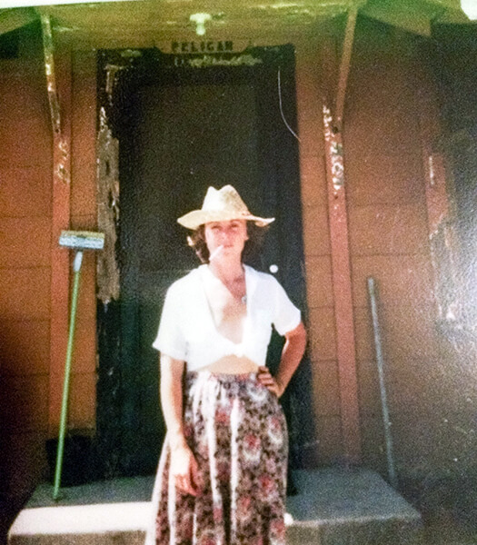 Rena Carney wears a straw hat and stands in front of her cottage door with hand on hip