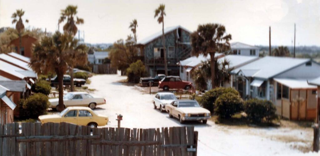 Two rows of small cottages withi cars parked in front of them and palm trees here and there