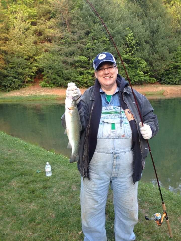 Lenny Lasater holding a large bass she caught in one hand and fishing rod in other standing in front of water