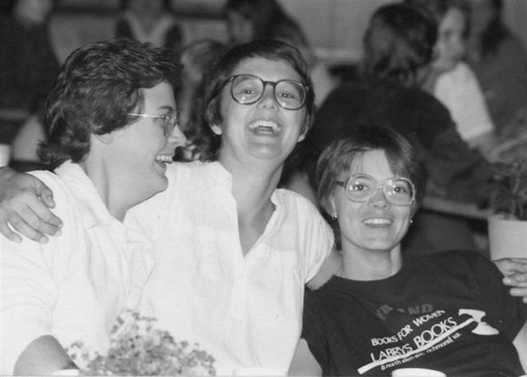 Three smiling women, the one in the middle with her arms around the others