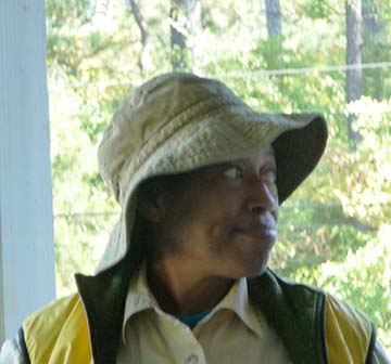 Blanche headshot, wearing a floppy hat and outdoor clothing, trees behind her.
