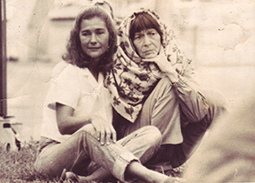 Bonnie and Barbara are seated on the ground outdoors, Barbara wrapped in a blanket.