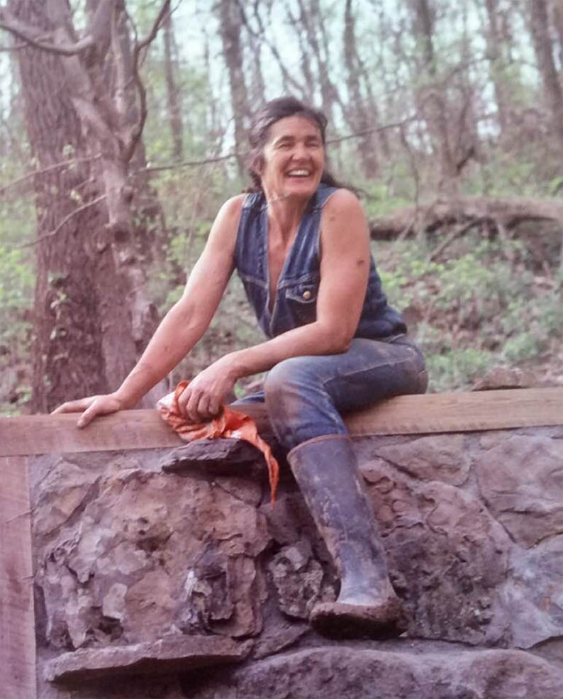 Diana Rivers in mud boots straddles a wooden rail on top of a stone wall