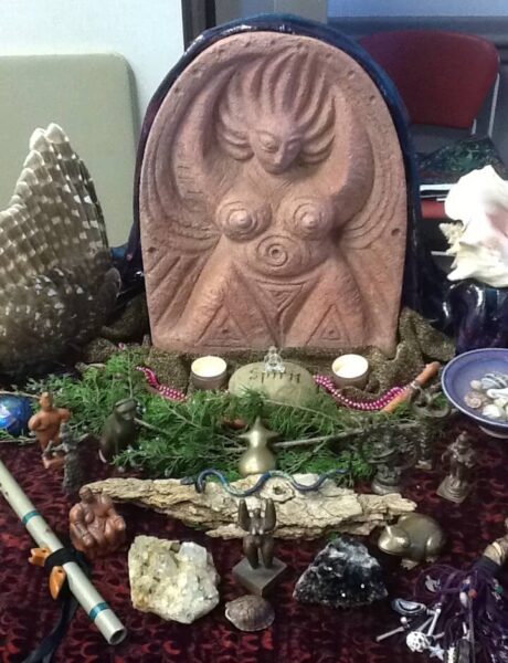 Clay sculpture of stylized goddess with rocks, crystals, small figurines, greenery, and other sacred objects in front of it