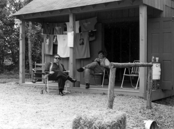 Beth Marschak and an unidentified woman are seated in folding chairs on the open front porch in front of a rustic cabin.