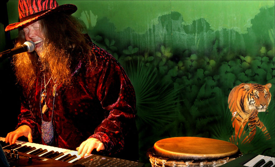 Flash Silvermoon wearing a tiger striped hat and velvet jacket, singing and playing keyboard, with a tiger backdrop.