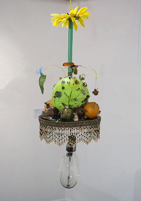 beaded stand above light bulb holding green globe covered in and surrounded by peculiar objects under tall sunflower