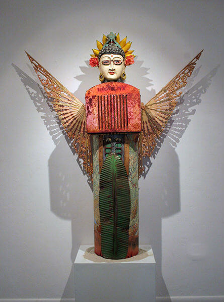 stylized winged buddhist goddess with lotus behind head in red and green bejeweled gown against a white wall carrying her shadow