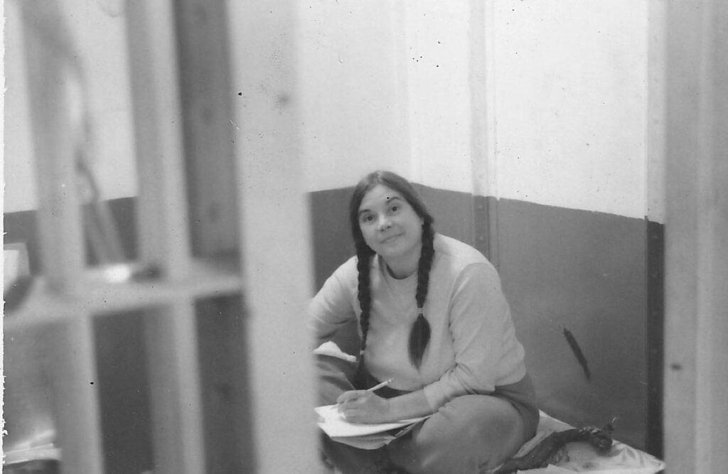 Corky Culver with long pigtails sits to write on her folded knees on a bunk in an open jail cell