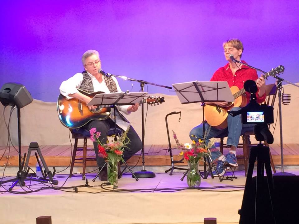 Ann and Jan in blue jeans, seated onstage in front of mics and music stands, with flower bouquets in front of them.