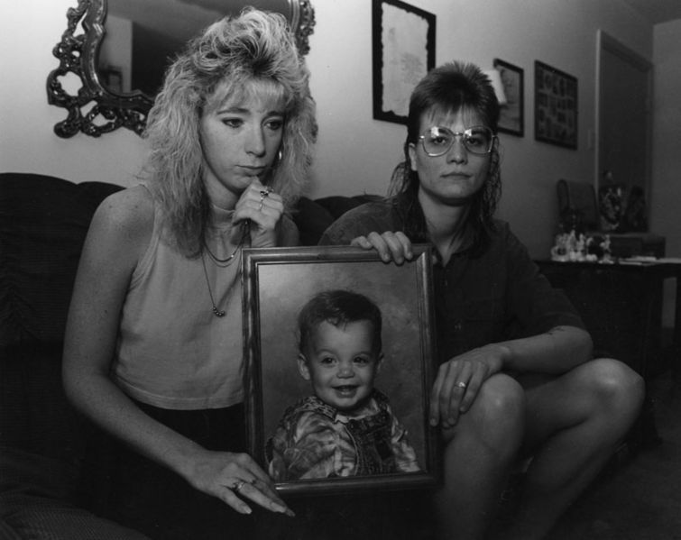 Two women looking sad, seated on a couch holding between them a large framed photo of a toddler boy.