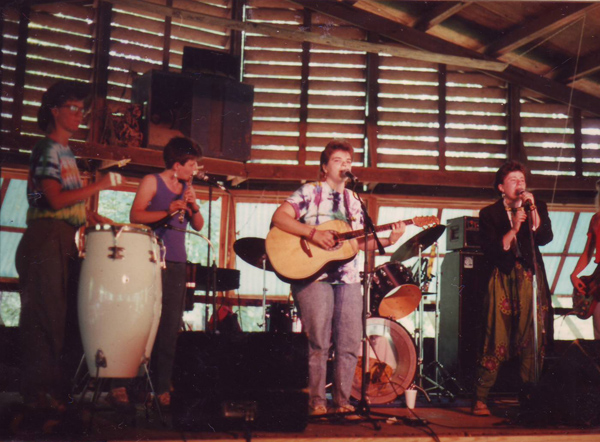 The band performing indoors. There is a big white conga drum on the left, and Carol Kraemer is playing guitar.