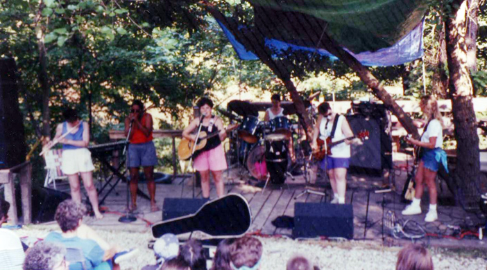 The band performing outdoors on a ground-level stage with trees behind them. Everybody is wearing shorts.
